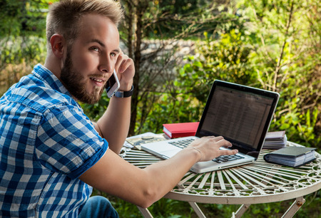 young man on a phone in front of his laptop outside.